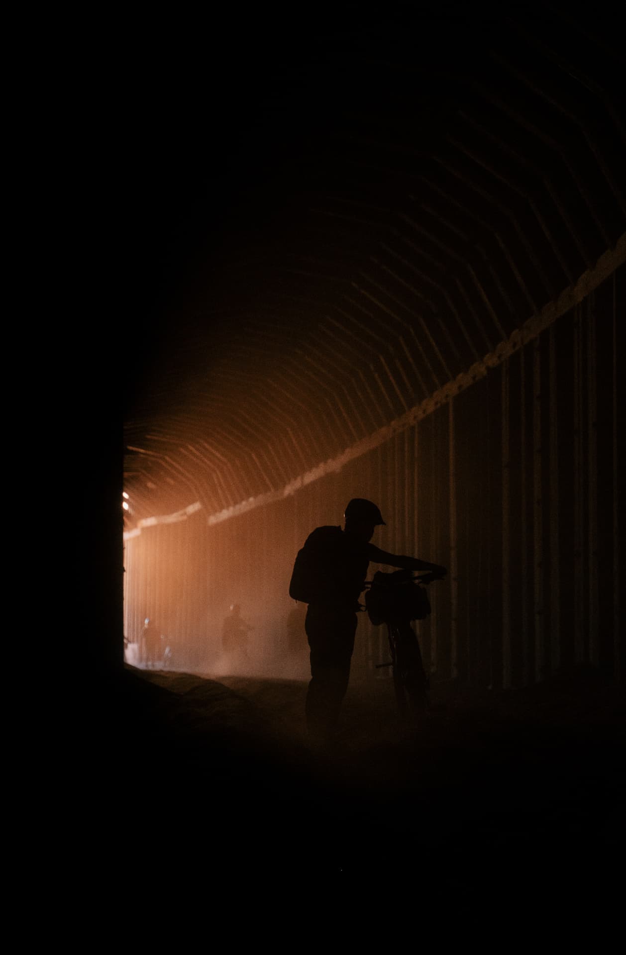 Silhouetted cyclist pushing a loaded bike through a dust-filled railway tunnel