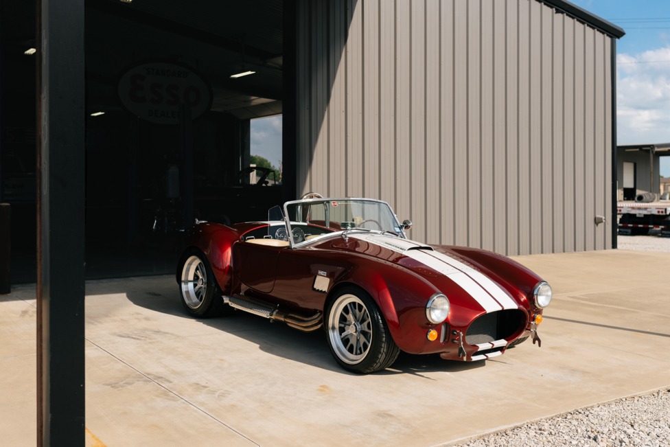 Deep red Cobra roadster parked beside a shop bay with a vintage Esso sign