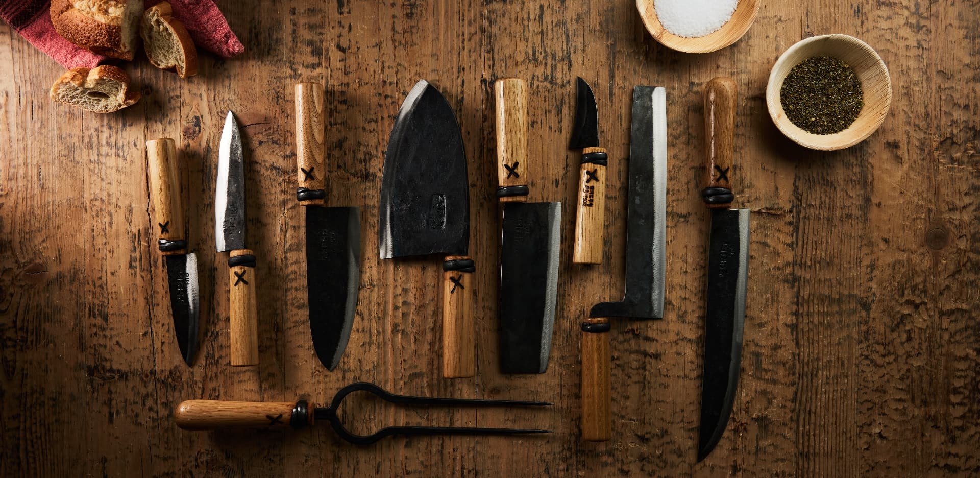 Overhead spread of hand-forged kitchen knives and a carving fork on weathered wood