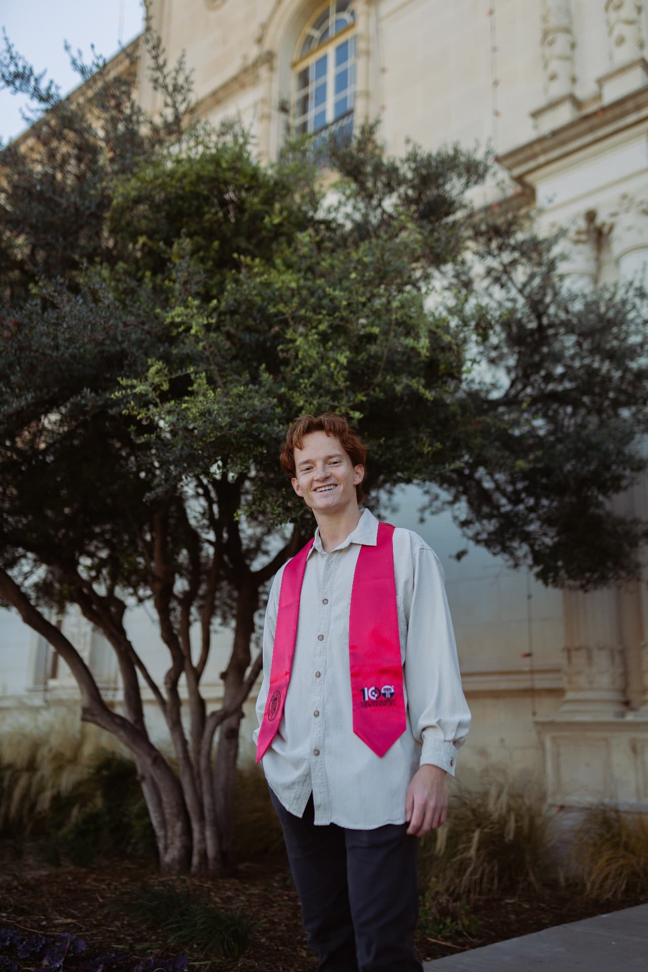 Smiling graduate in a pink stole standing beneath an olive tree