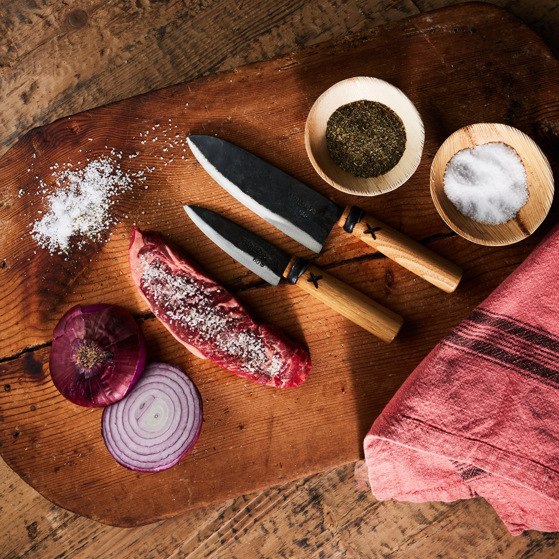Overhead still life of forged knives, seasoned steak, salt, and red onion on a wood board
